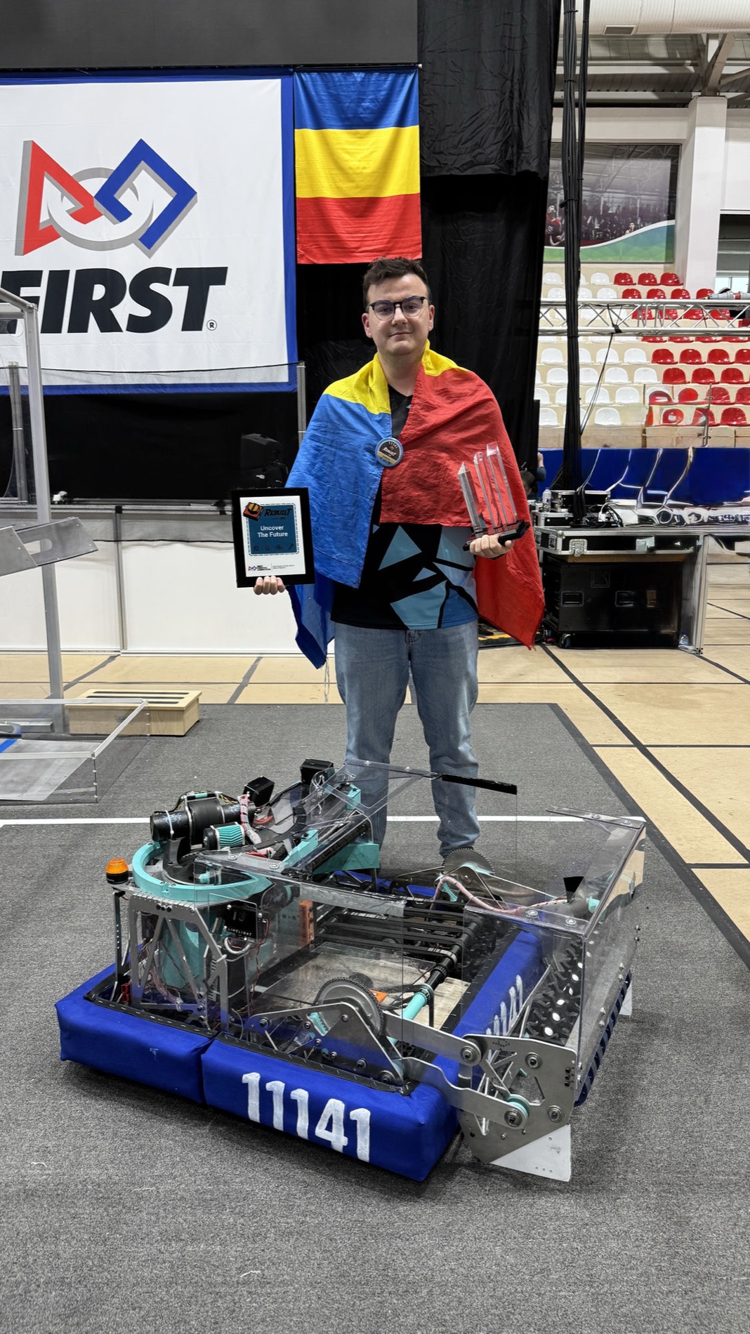 Holding the Rookie All-Star award and trophy behind Team AlphaZ robot #11141, draped in the Romanian flag
